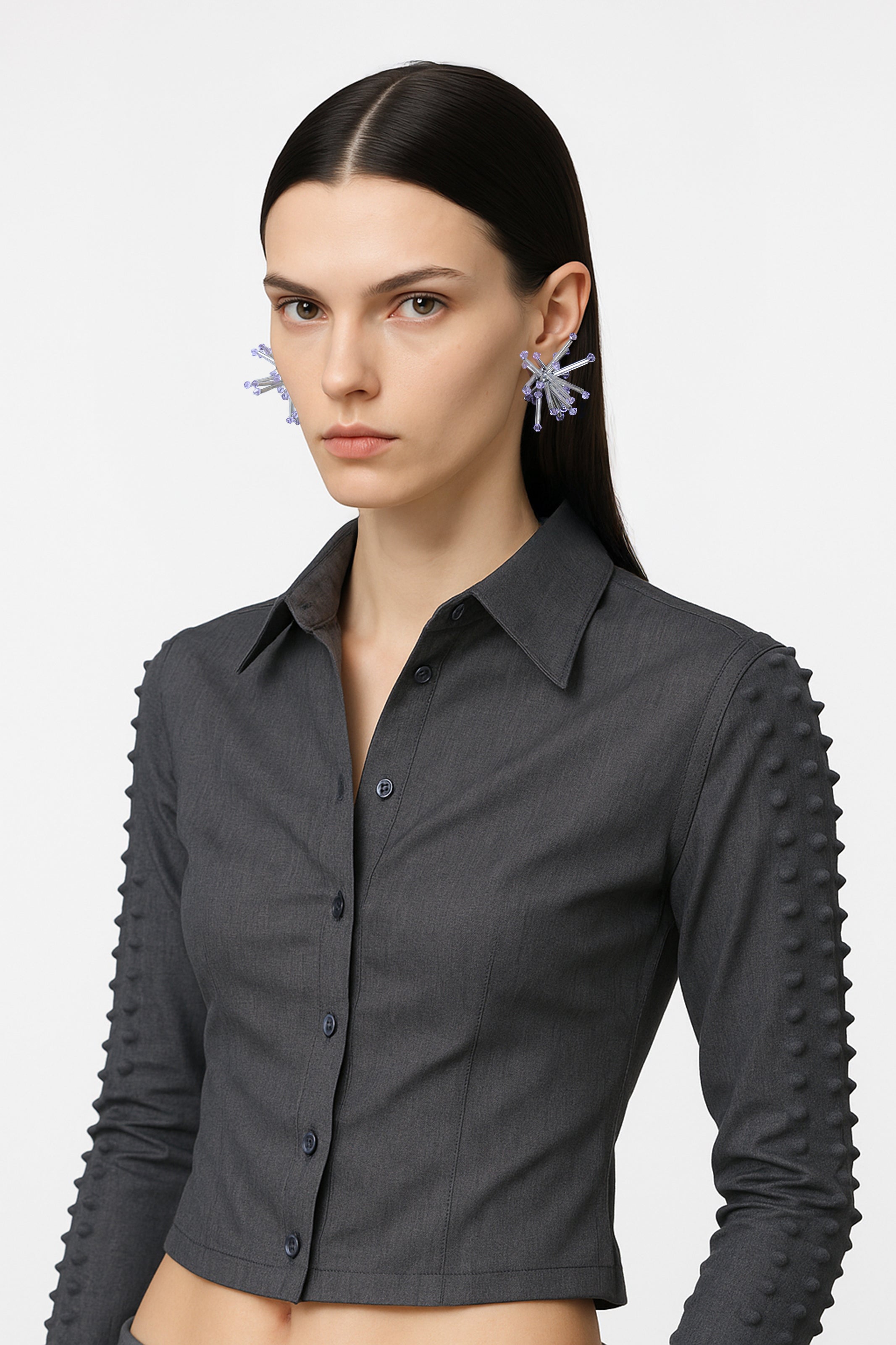 A silver small cluster floral inspired pair of earrings with violet crystal bicones tips worn by an AI model with grey spiky jacket and dark hair, view from 3/4