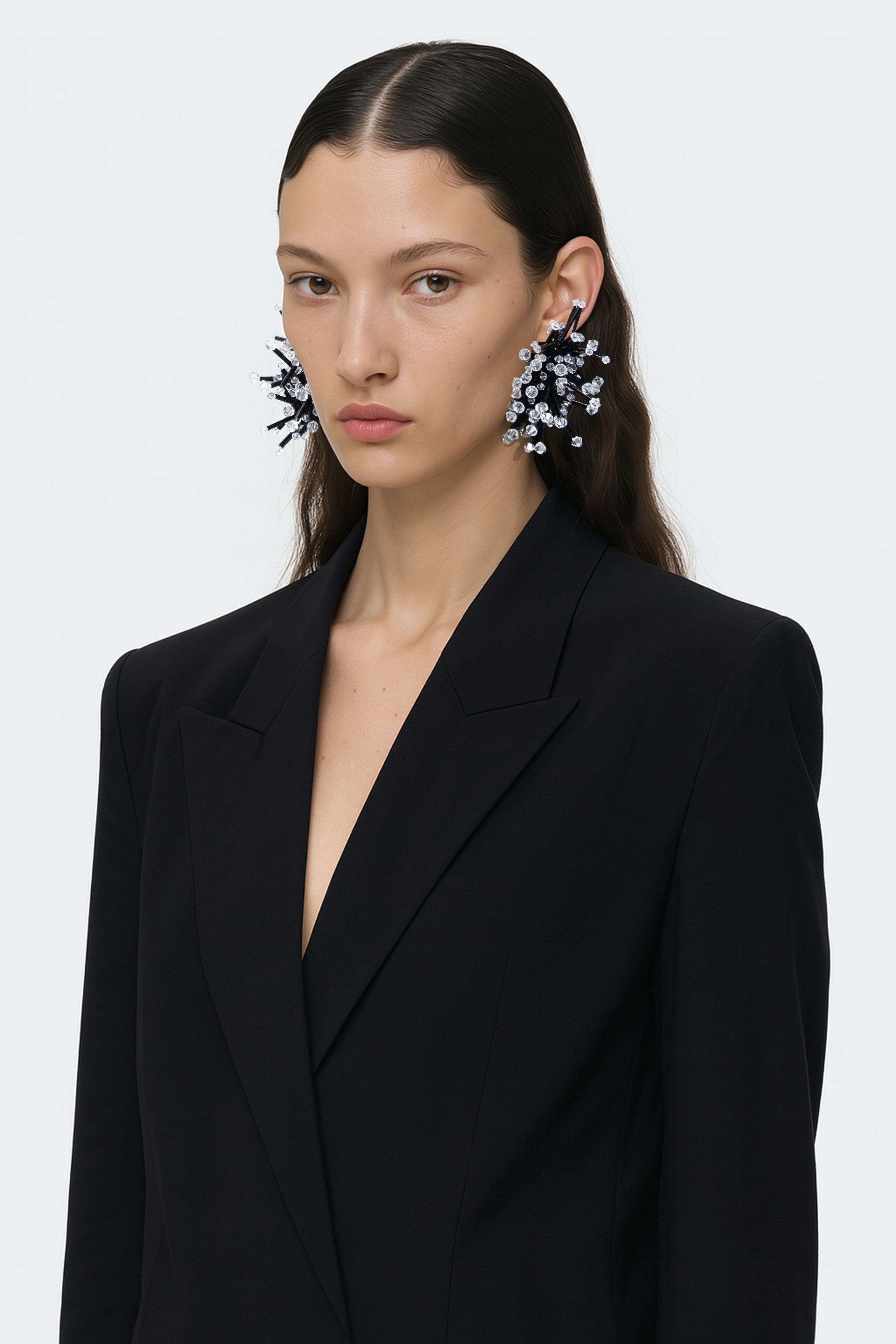 A black cluster floral inspired pair of earrings with clear crystal bicones tips worn by an AI model with black tailored jacket and brown hair view from 3/4