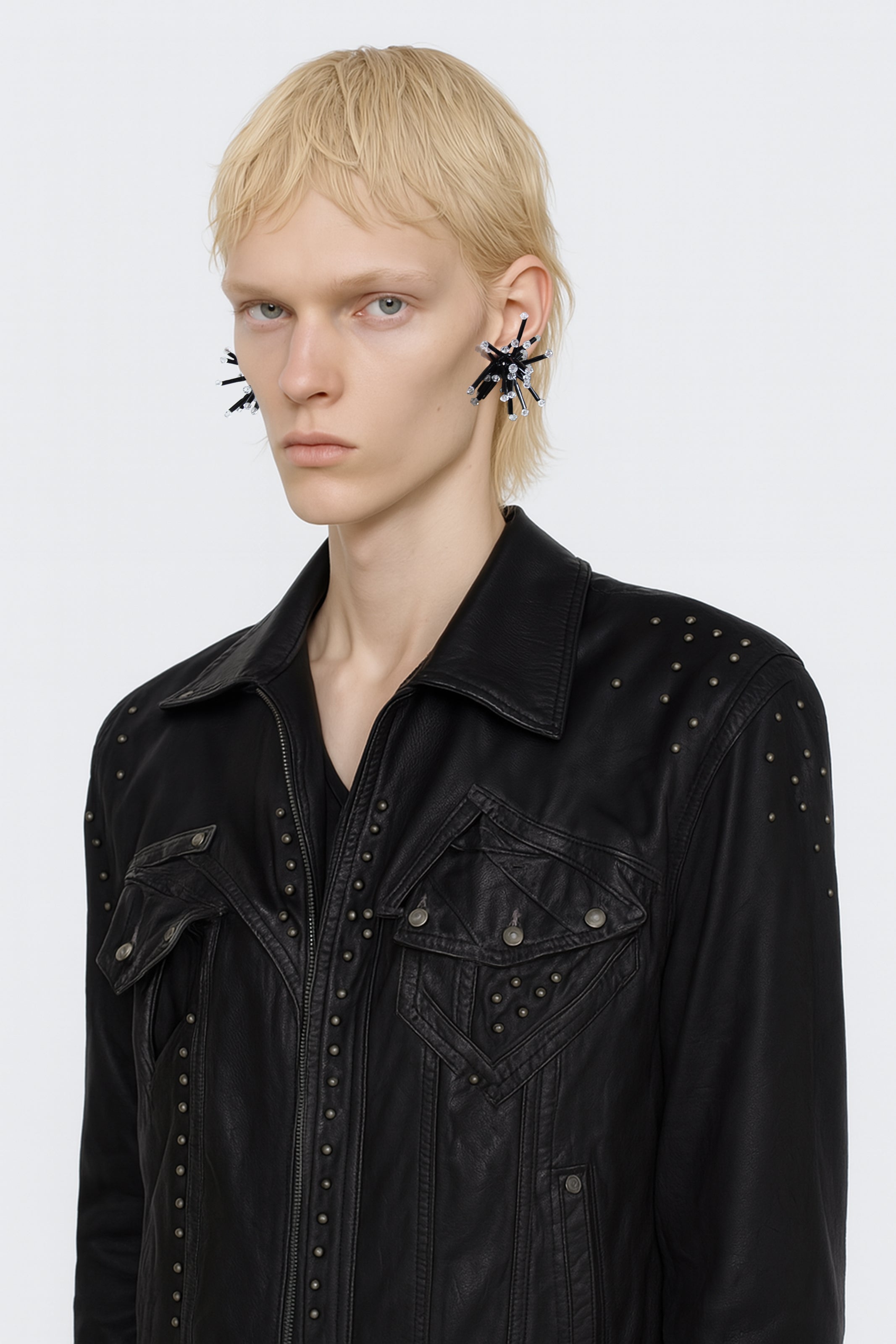A black small cluster floral inspired pair of earrings with clear crystal bicones tips worn by an AI male model with leather black jacket and polar blond hair view from 3/4