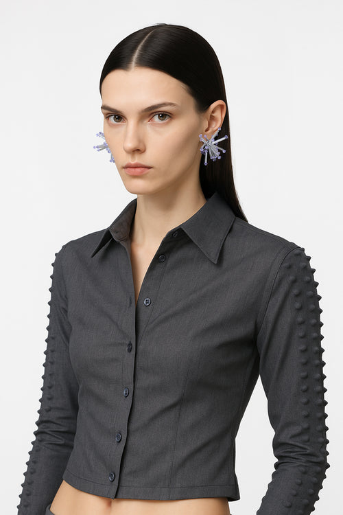 A silver small cluster floral inspired pair of earrings with violet crystal bicones tips worn by an AI model with grey spiky jacket and dark hair, view from 3/4