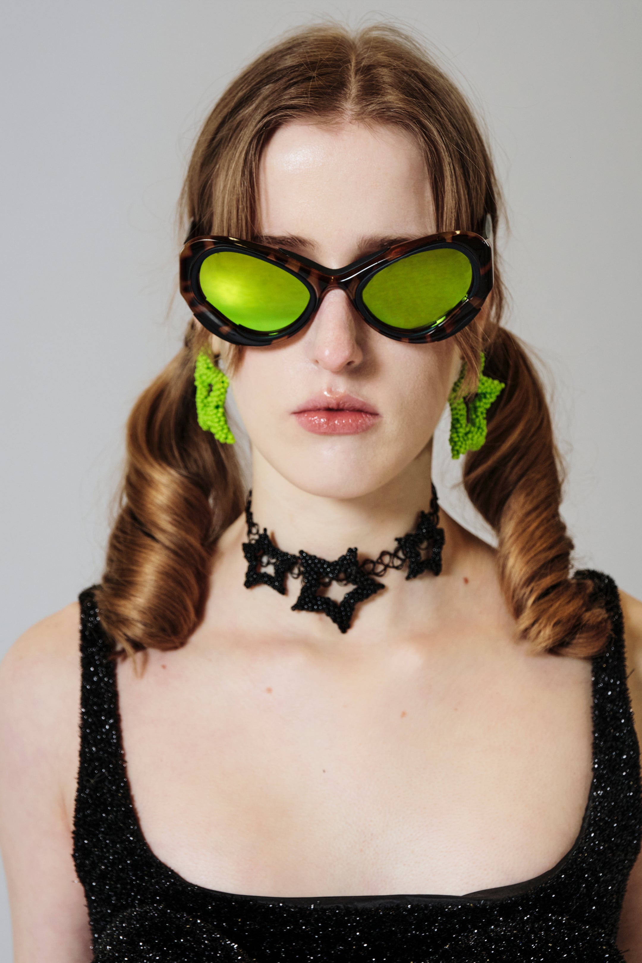 Model wearing a black outfit with green beaded star earrings against a gray background