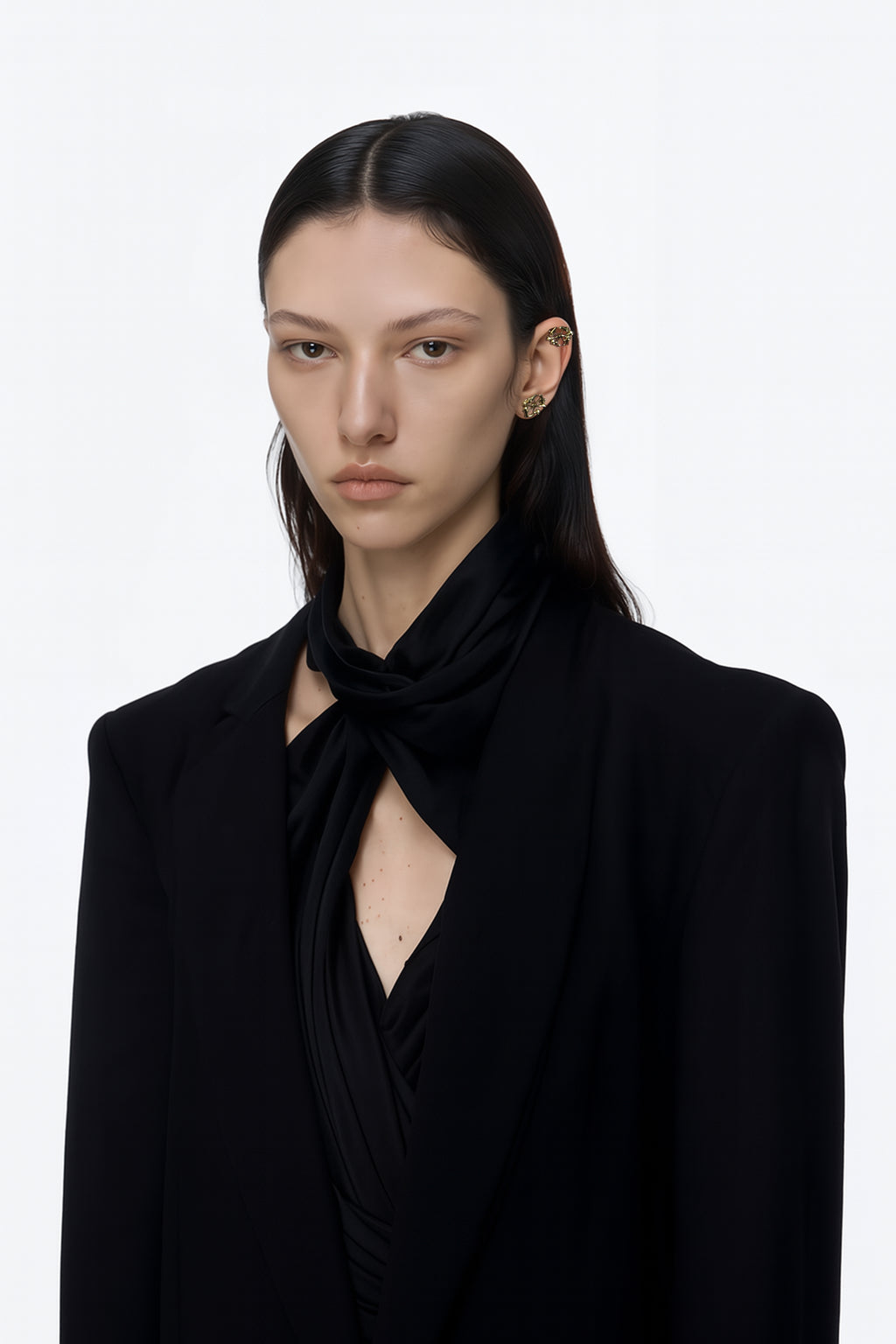 A brass gold pair of stud logo earrings with floral inspired spikes pair of earrings  worn by an AI model with black tailored jacket black scarf shirt and dark long hair view from 3/4
