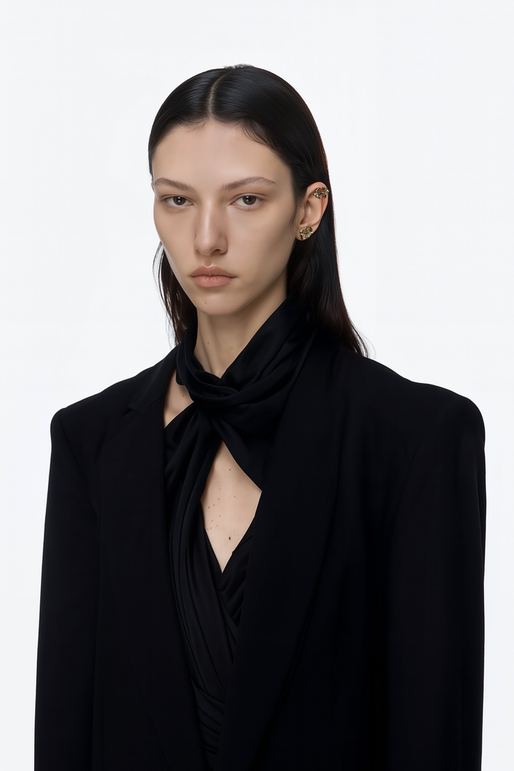 A brass gold pair of stud logo earrings with floral inspired spikes pair of earrings  worn by an AI model with black tailored jacket black scarf shirt and dark long hair view from 3/4