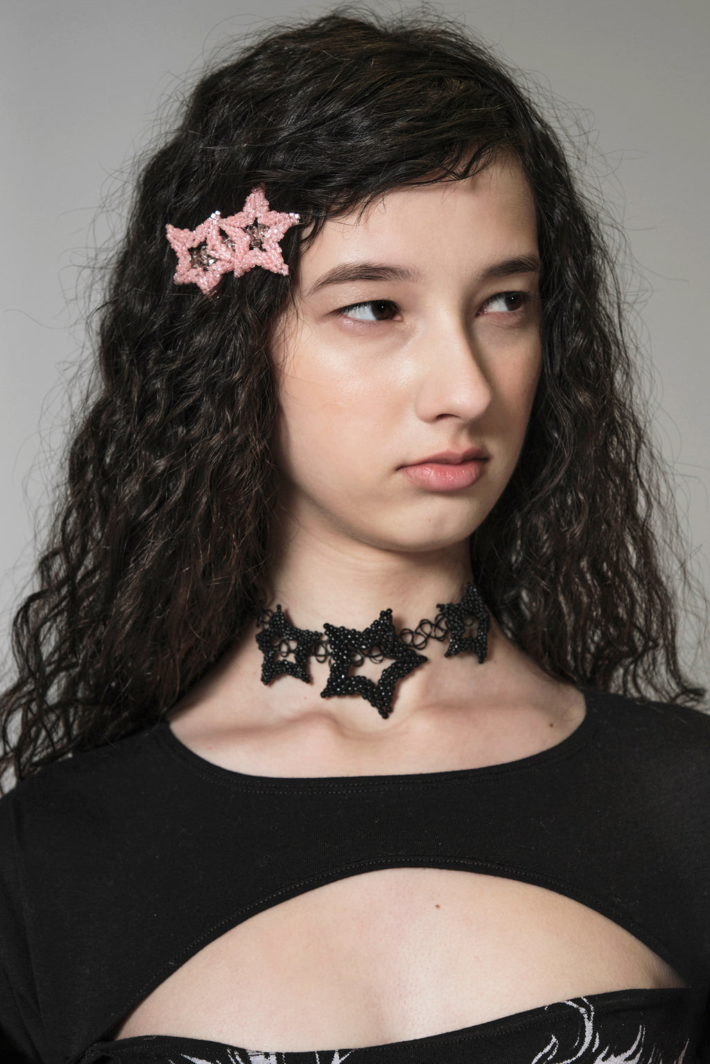 Woman wearing a pink star beaded hairclip and a black star-shaped choker with a gray background