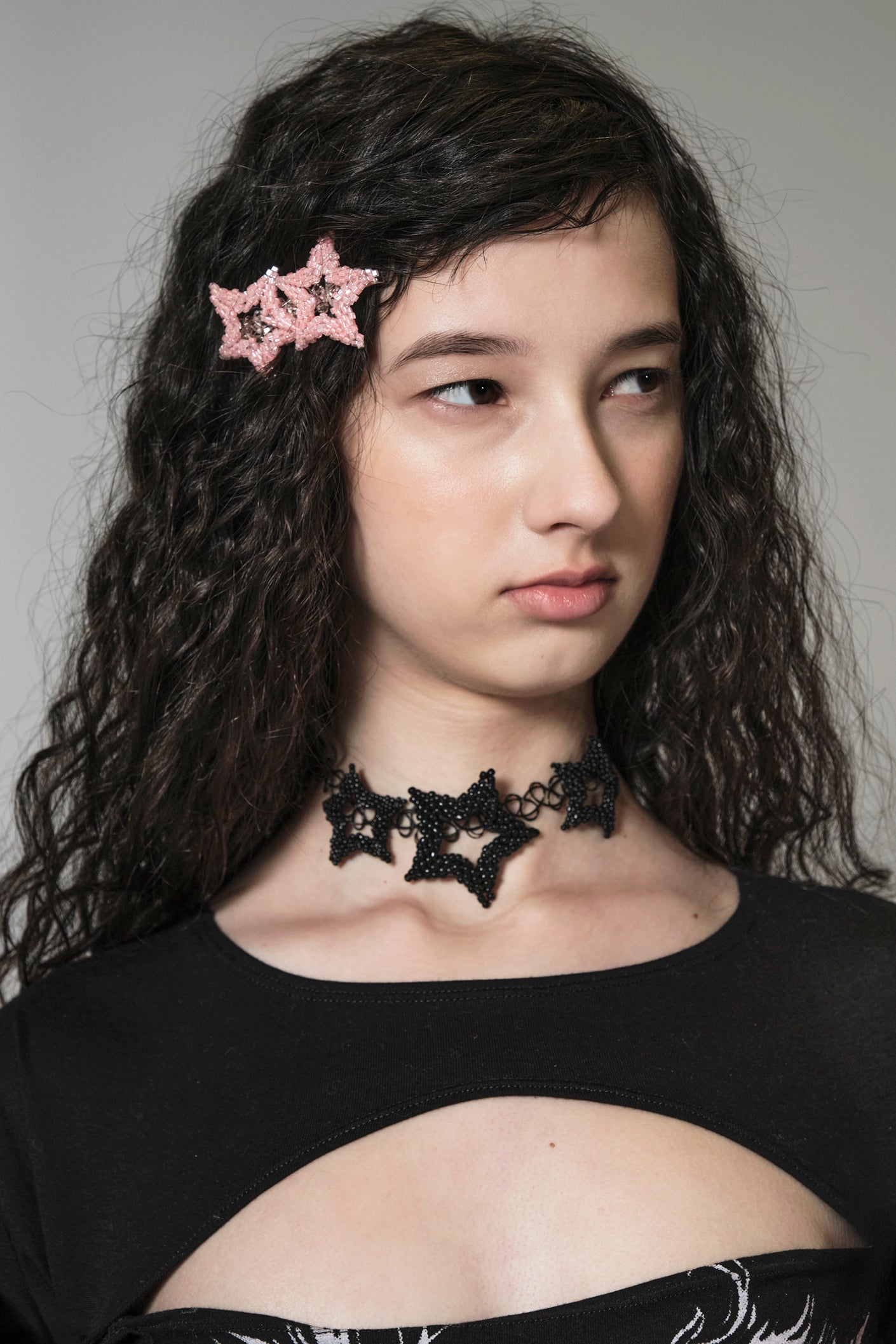 Woman wearing a pink star beaded hairclip and a black star-shaped choker with a gray background